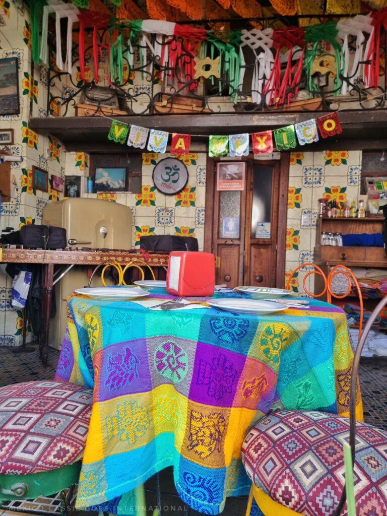 interior of a colourful restaurant - one table in foreground with brightly coloured tablecloth, 2 chairs and table settings. in background bunting that says viva mexico