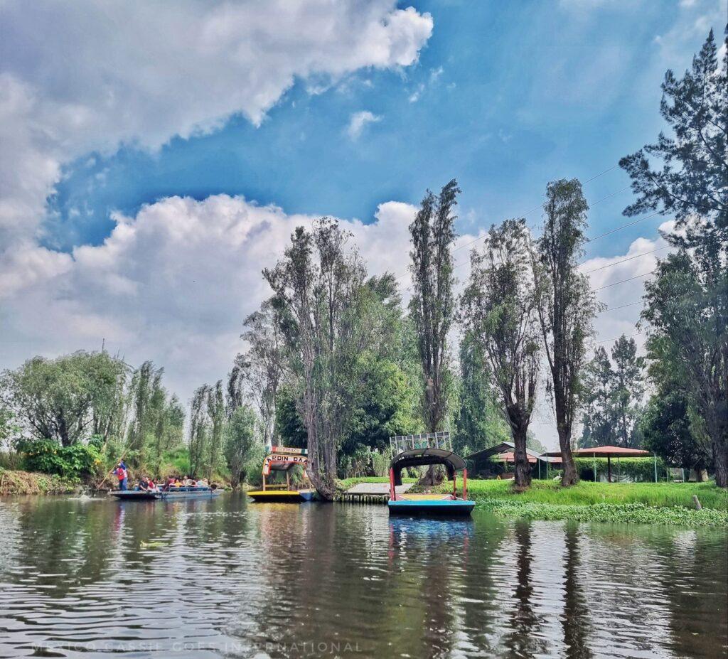 calm river/canal with a few trajinera long boats on it, trees and blue cloudy sky