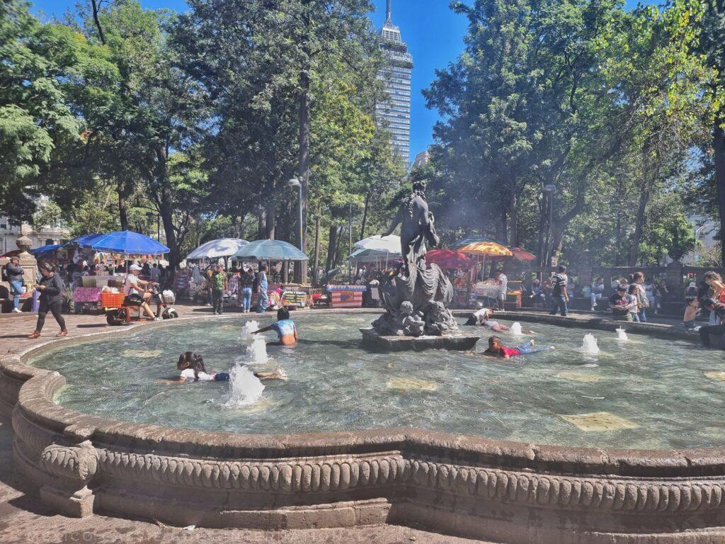 kids playing in water in fountain in a park, trees all around