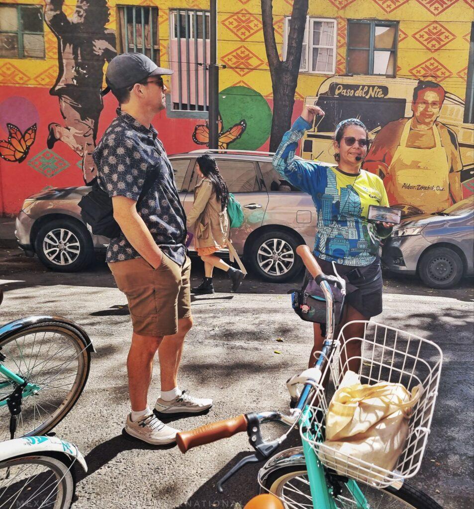 Gabi from Poráy talking to a group of riders on her bike tour. in shot only her and 1 guy in shorts, handlebars of green bike in foreground