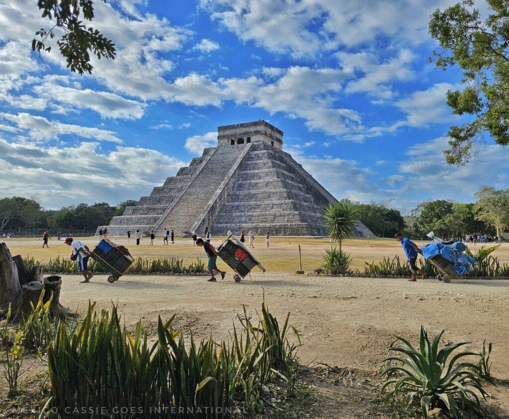 chichen itza main temple against blue cloudy sky, 3 people carrying loads walking across path
