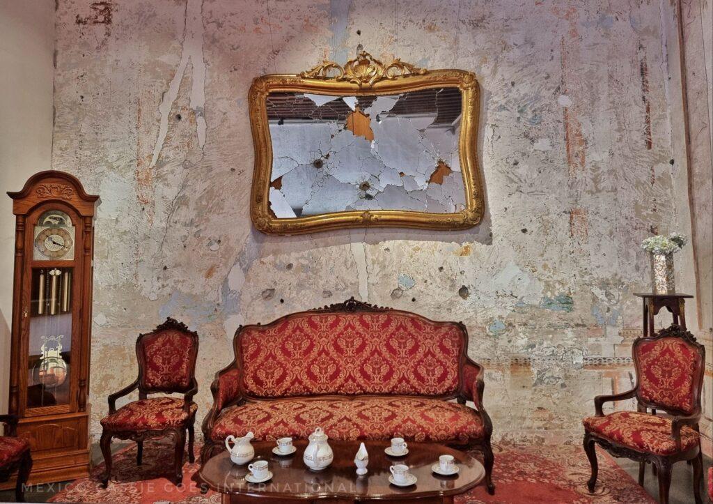 old fashioned sofa and 2 chairs with red pattern, coffee table with tea cups and tea pot, grandfather clock and shattered mirror above the sofa