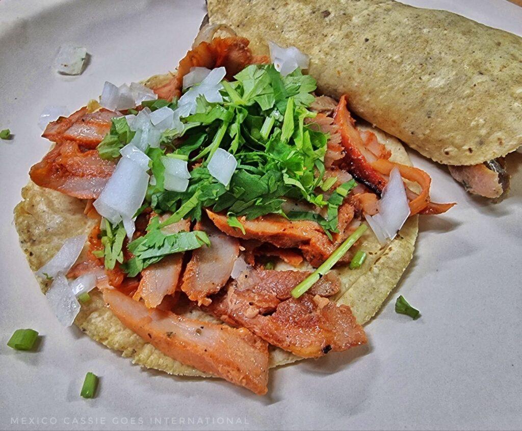 one open taco and one that's wrapped up on itself - slightly reddish pork (because it's seasoned) with cilantro and chopped onion on top