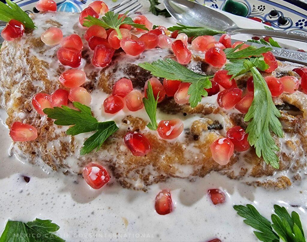 chile en nogada on a plate - battered chile pepper in white sauce with parsley and red pomegranate seeds - you can just see the knife, spoon and fork behind