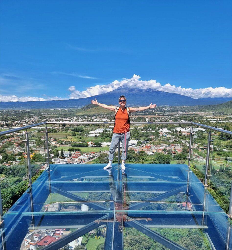 Cass standing, arms open, on glass platform with Popcatépetl volcano behind