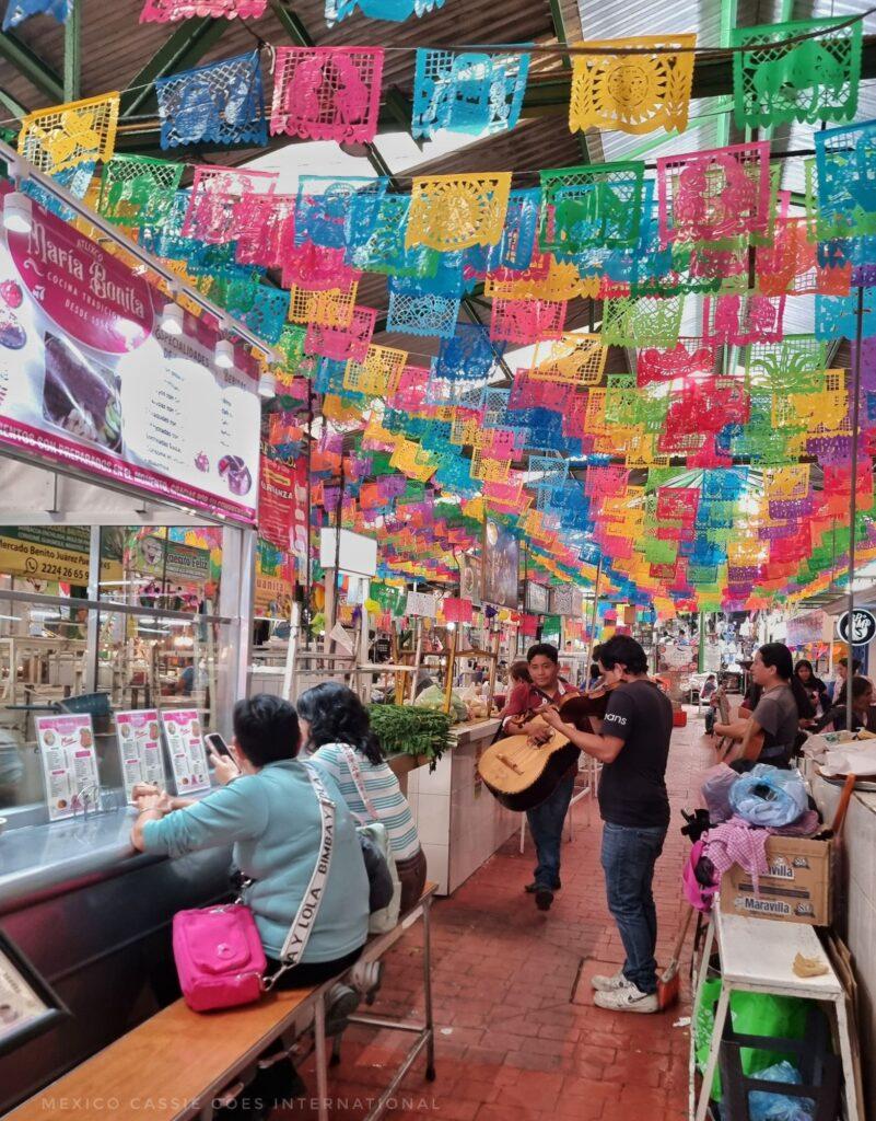 colourful market scene, bunting, people sitting on benches to eat, 2 people with a guitar