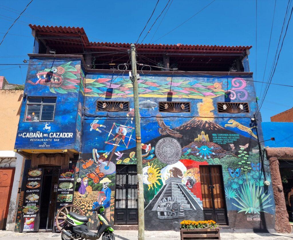 3 storey building with street art mural over it - scenes of day of the dead, a pyramid, volcano and traditional depictions