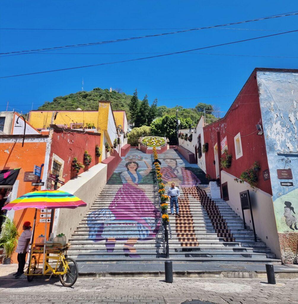 view looking up a large flight of outdoor stairs that are painted with a picture of a man and woman in traditional Mexican dress. one man standing on steps, ice cream selling with rainbow parasol at base of steps