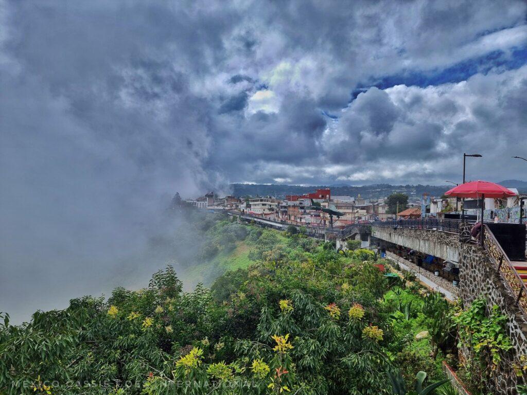 view along a barranca with trees below and cloud to left, town visible on right of screen