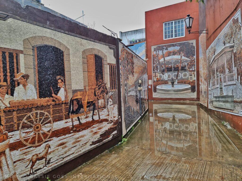 view down callejón de los huesos, murals on both sides of street, one at end of the kiosk, also reflected in puddle