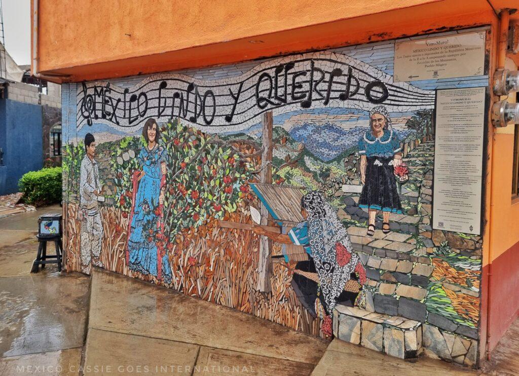 mural that reads "México Lindo y Querido" written along musical staves, picture underneath is nature scene that includes 3 women and a man in the fields