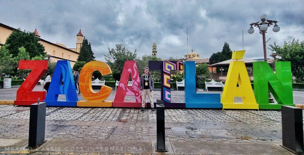 Cass standing in front of the Zacatlan letters on the plaza on a grey day