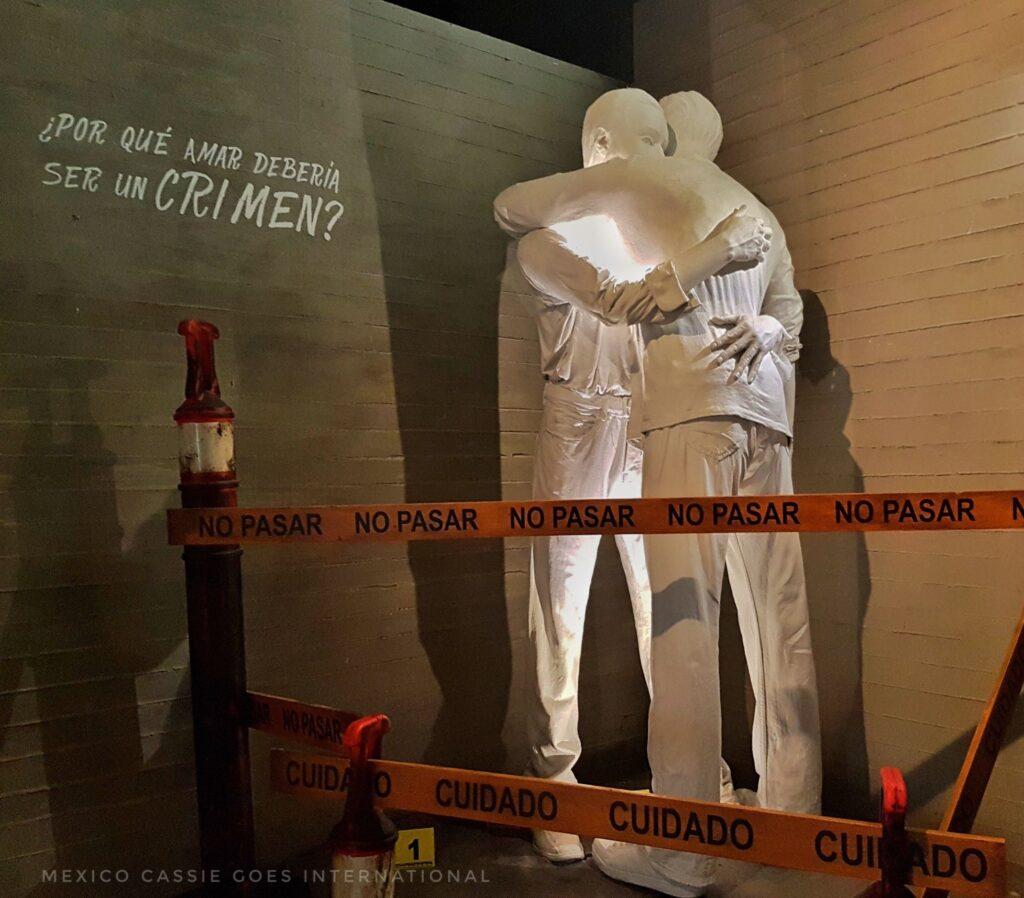 sculpture of 2 people hugging. on wall next to them it says "por qué amar deberaia ser un crimen?". yellow police tape around sculpture that reads "no pasar, cuidado"