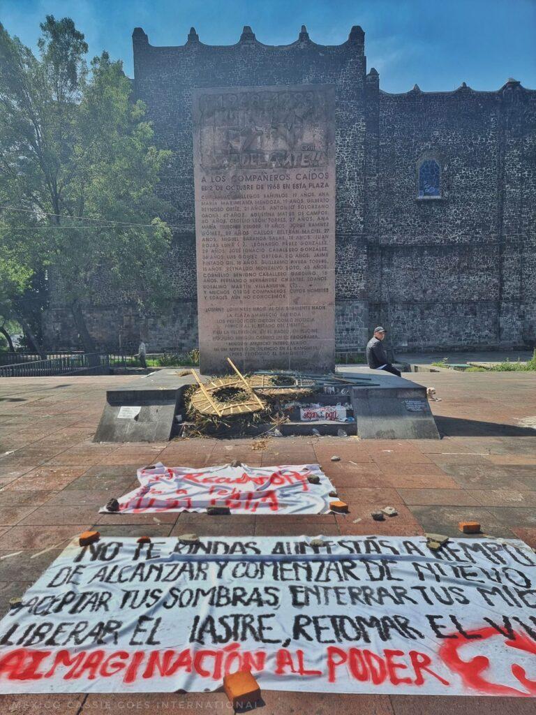 photo of the memorial stone for the students massacred in 1968 - white sheet with red and black writing in foreground, man sitting by the memorial stone