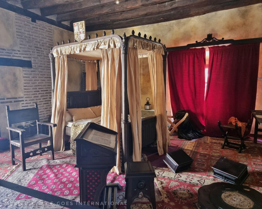 medieval room with 4 poster bed, red capret and curtains and wooden chair by bed. bed has white curtains