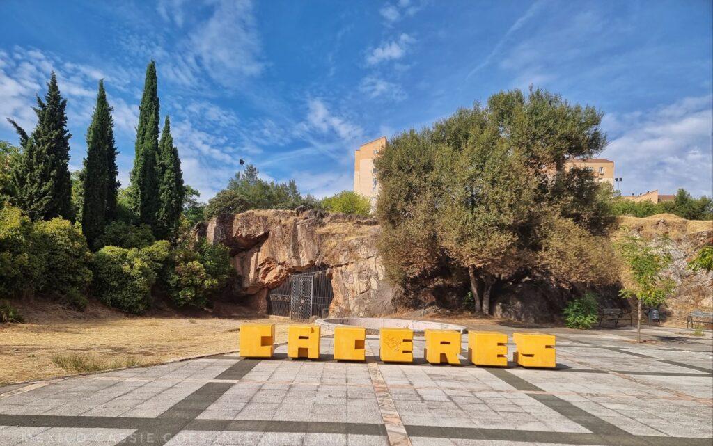 small yellow block letters saying "Cáceres" on a plaza in front of a cave, trees behind too