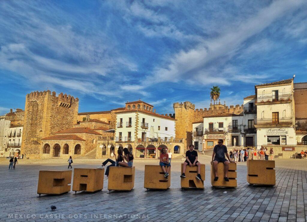 Caceres block letters with people sitting on top of them. large plaza with old buildings all around, blue sky