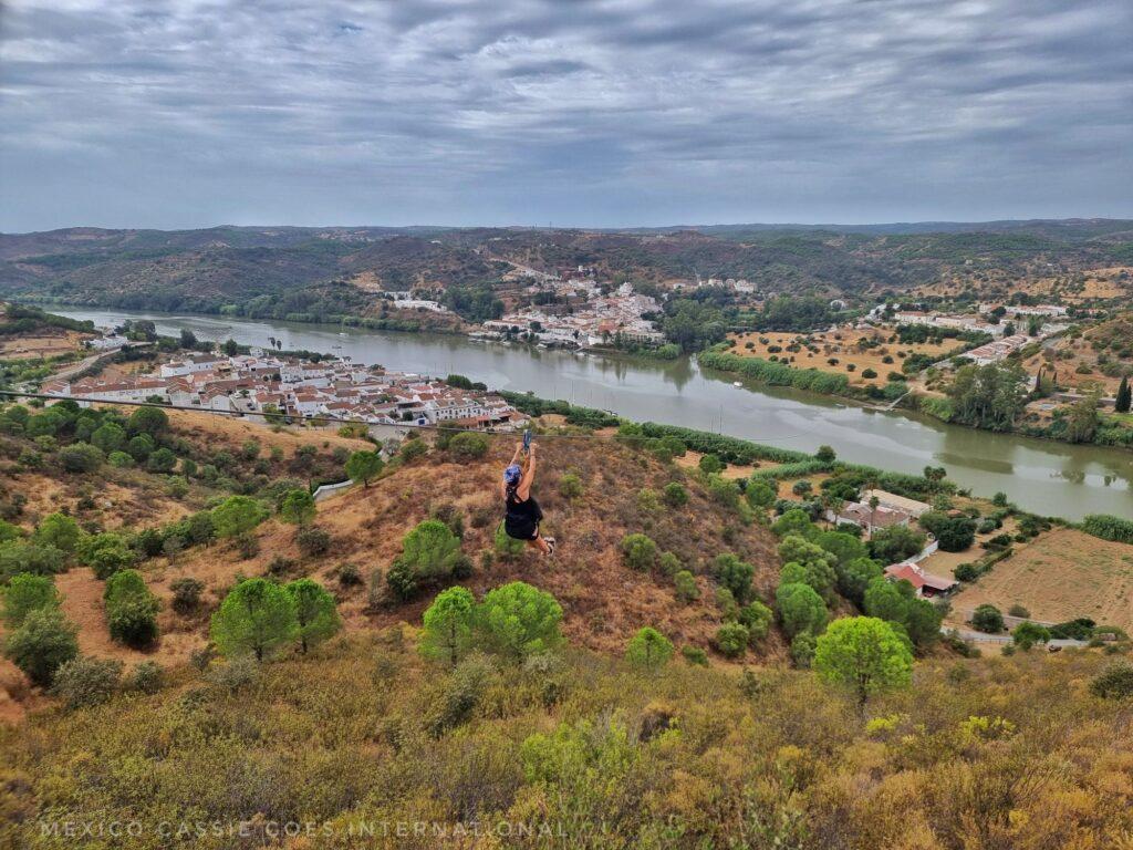 person ziplining over fields, trees and a river on a cloudy day