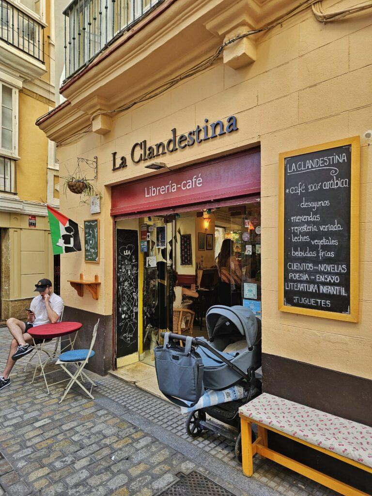 coffee shop La Clandestina from outside, yellow walls, pram and man sitting gat table, chalk board