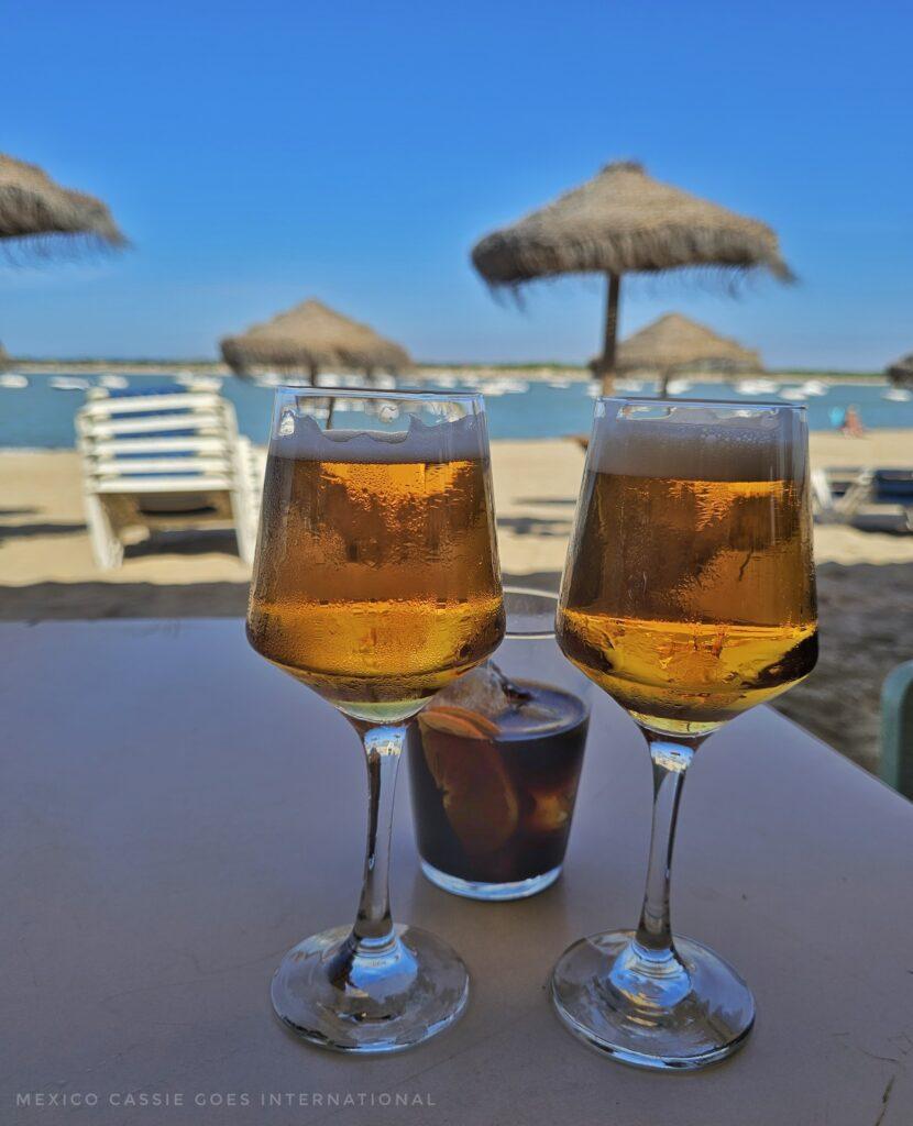 2 beers and a vermouth on the table, palapas, beach and sea behind