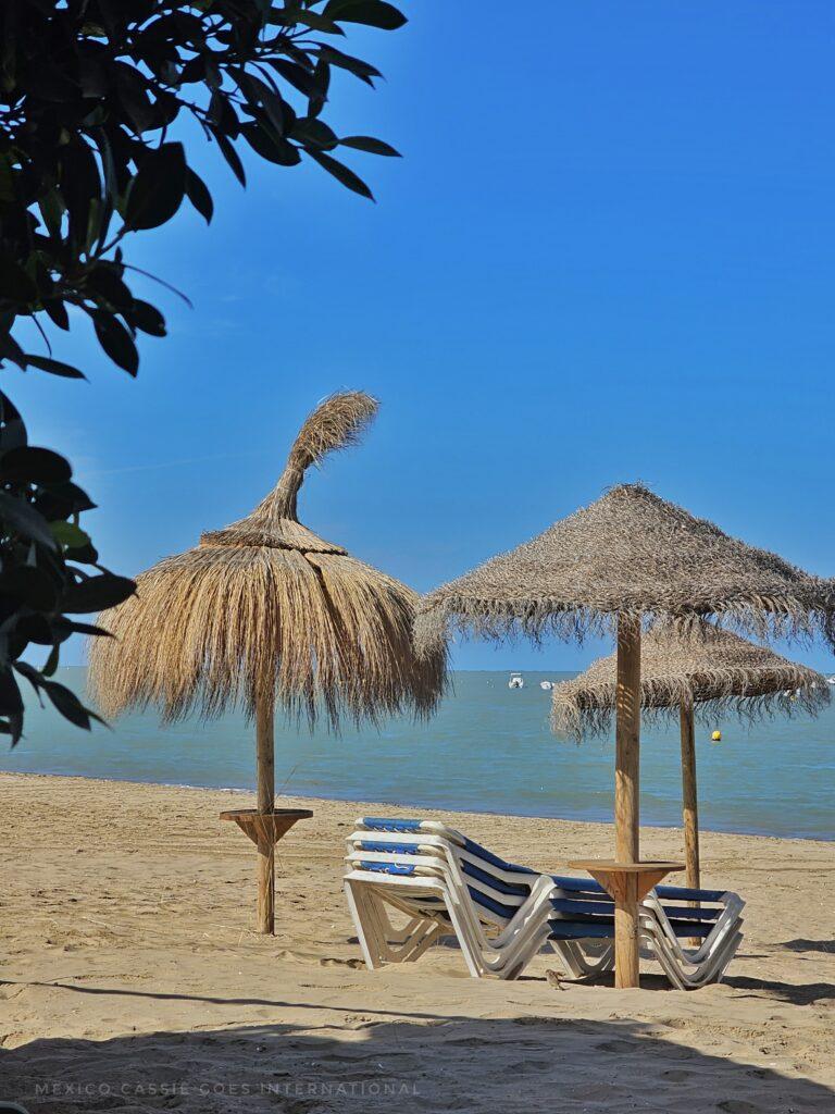 3 palapas on a beach, pile of sun loungers, blue sky, sand, tree in corner of shot