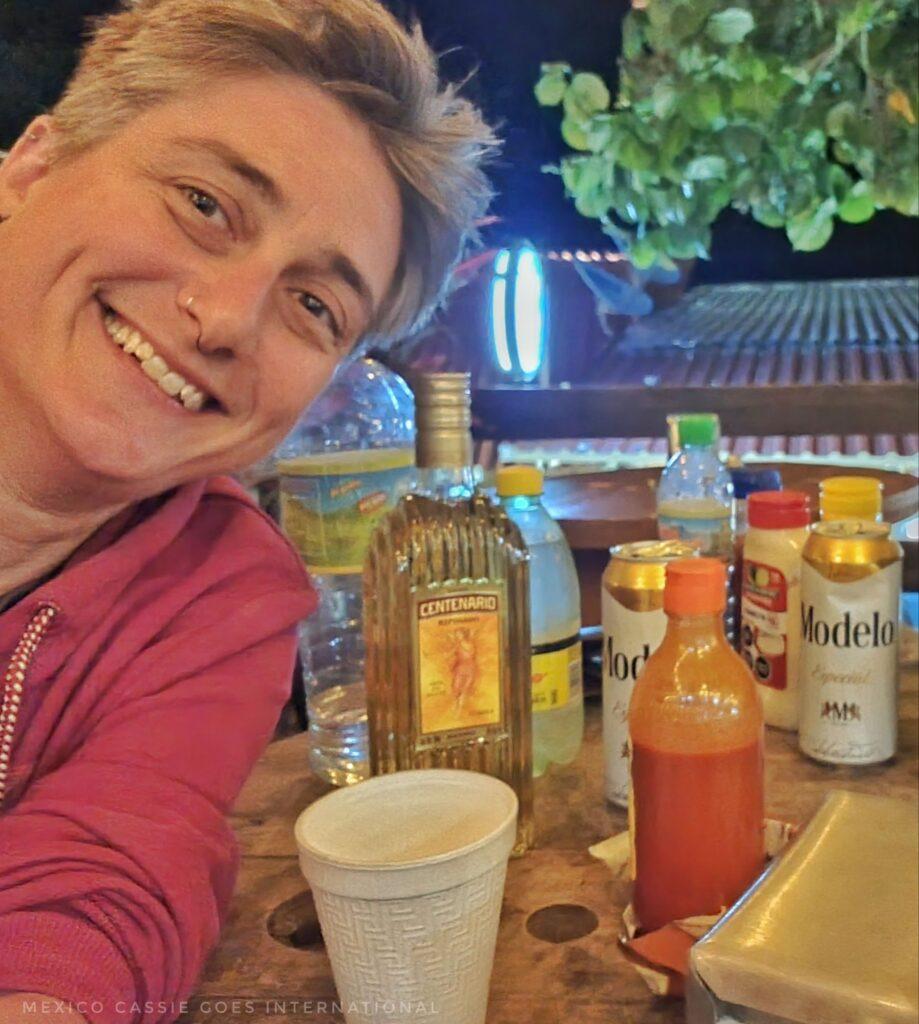 Cass putting head into shot - bottle of tequila on table, plus beers, water bottle and plastic glass
