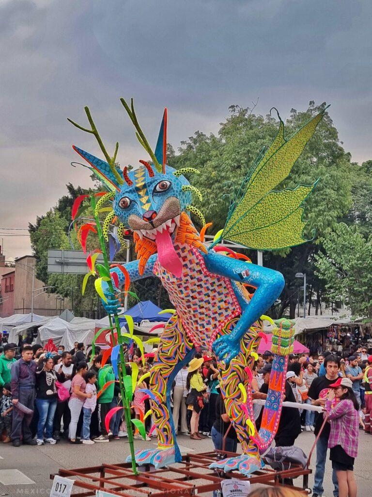 alebrije looking right at camera - crowds of people and trees behind. alebrije is mostly bright blue with wings and two arms, ears and horns