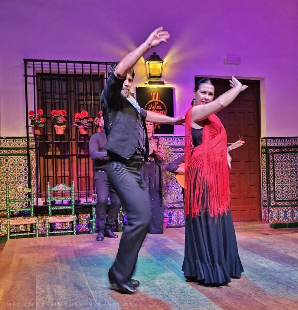 man and woman dancing flamenco on wooden stage. woman in black dress with red scarf. man in black. both with right arm raised