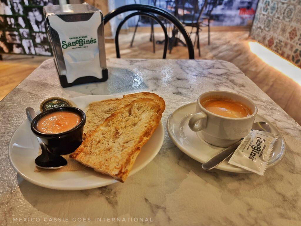 coffee in a white cup, plate on left with slice of brad and bowl of tomato. napkin holder and chair behind table