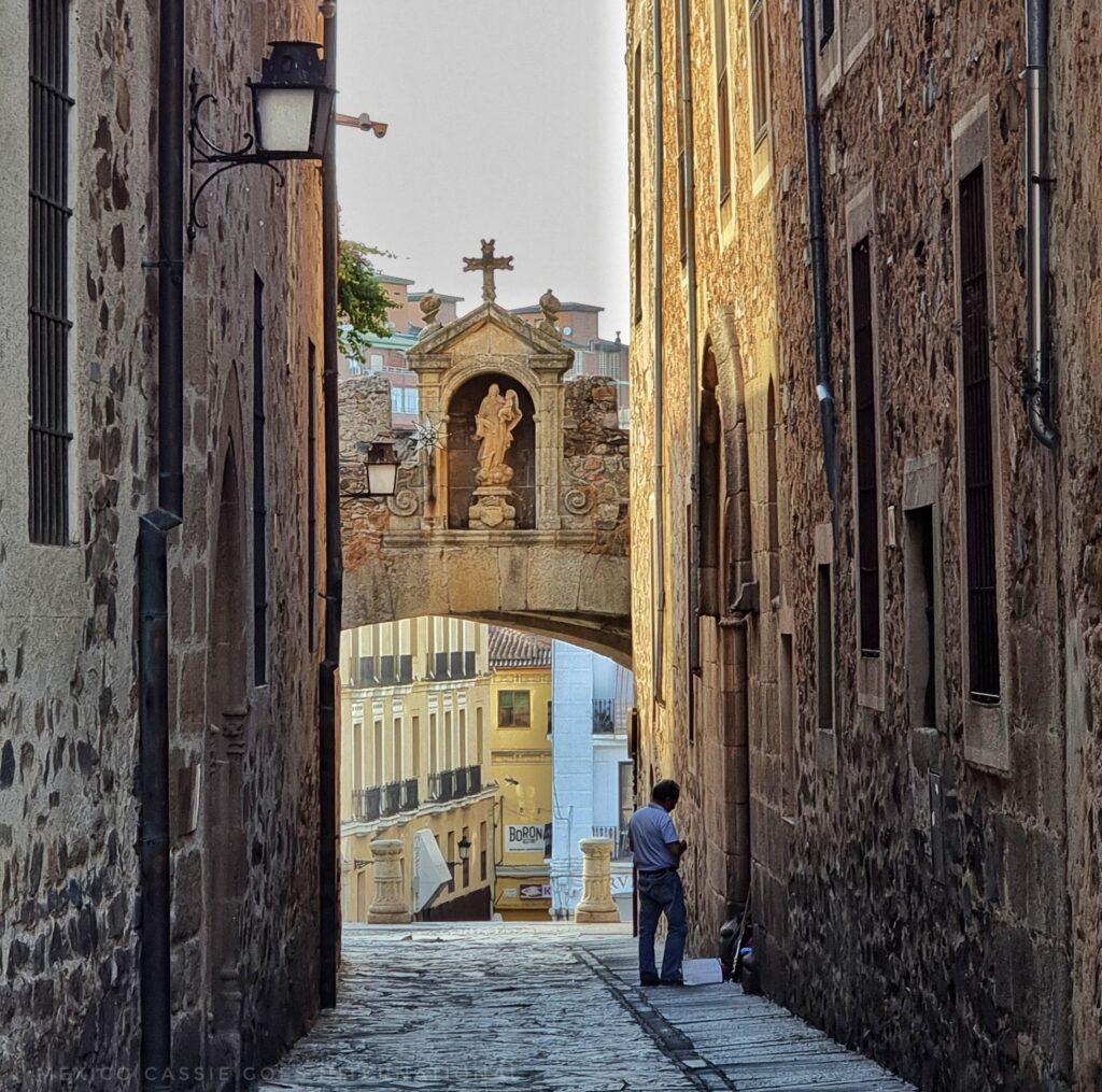 extremely narrow road between two buildings, arch over road, one person standing on right