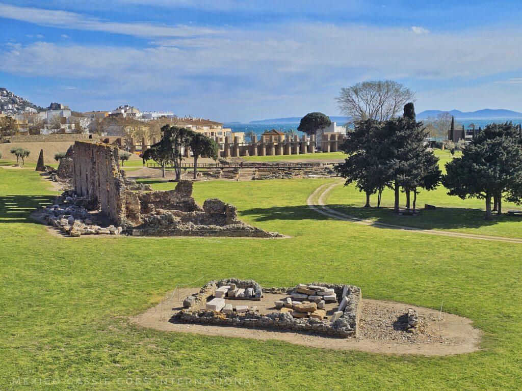 view over archaeological site that's mainly grass, modern town in background, trees and blue sky