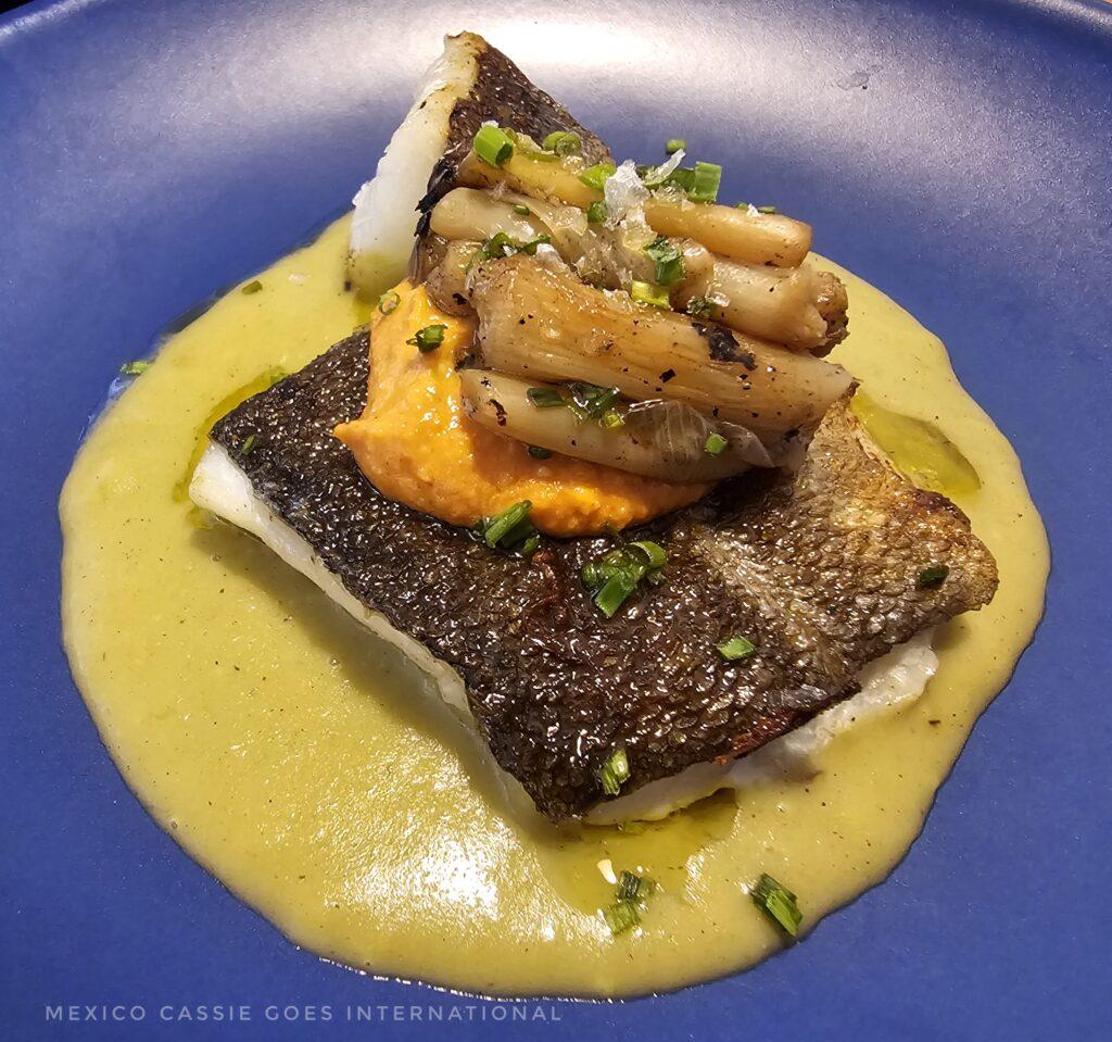 blue plate with piece of fish in a yellow sauce with veg balanced on top (sounds gross, looks great)