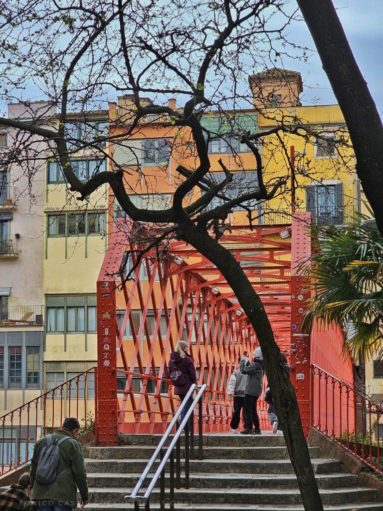 entrance way to red iron work bridge, few steps up and colourful houses behind. tree in foreground
