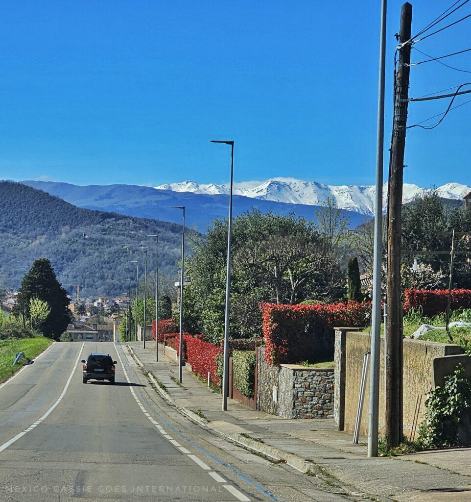road with one car in front, snow capped mountains in background, tree covered hills in front
