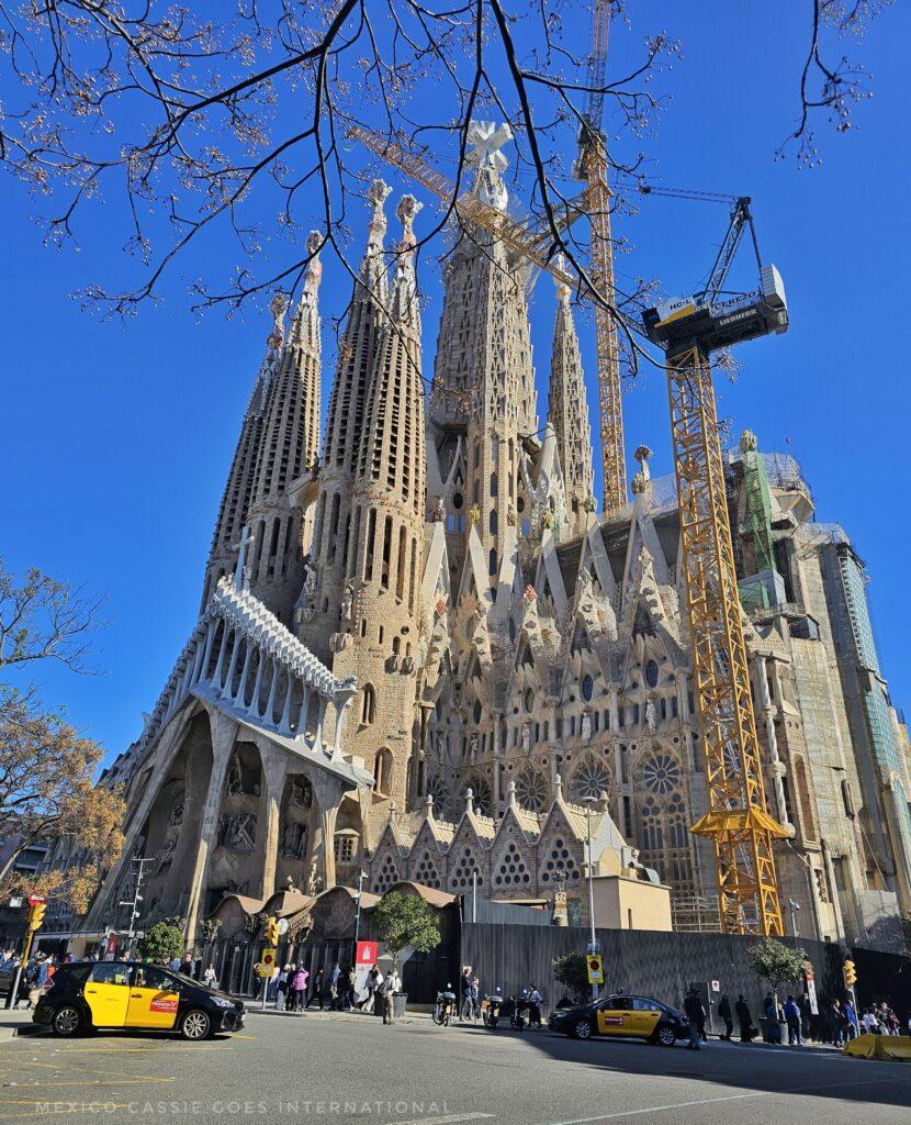 photo of Sagrada Familia, yellow crane on right, people milling around, church in full