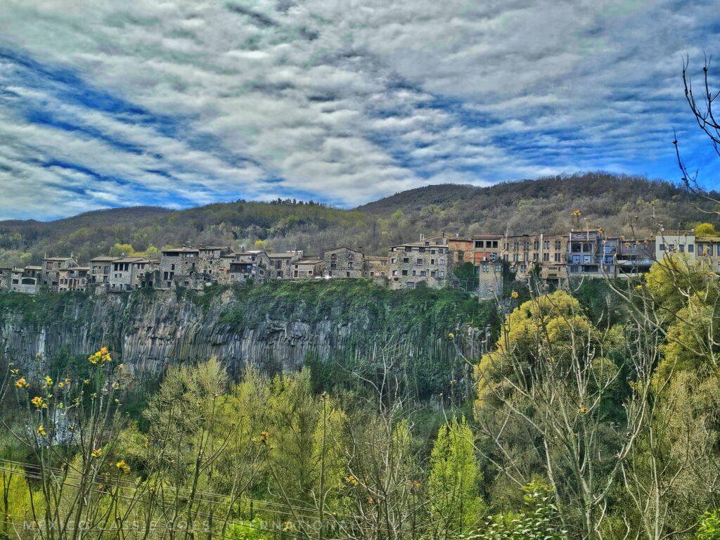 view of a medieval town on a cliff face, blue sky, white clouds, green trees in front