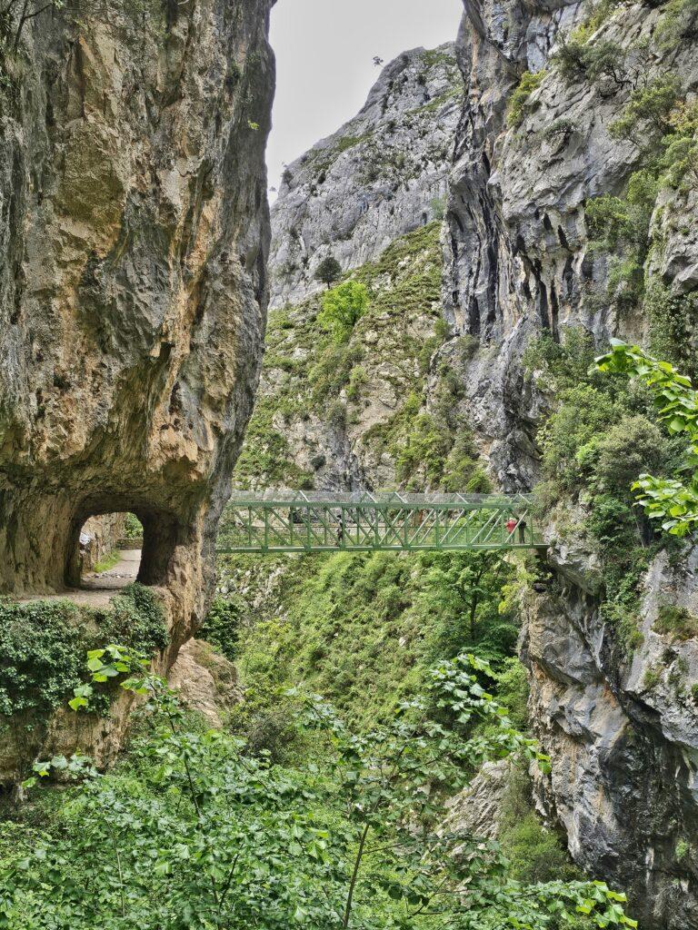 green metal bridge between two narrow mountain paths over a gorge - on left there's a tunnel visible through the rock