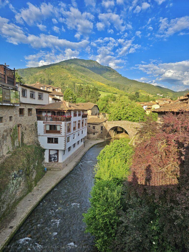 small river with bridge at far end of photo, old buildings on either side and green hills in distance