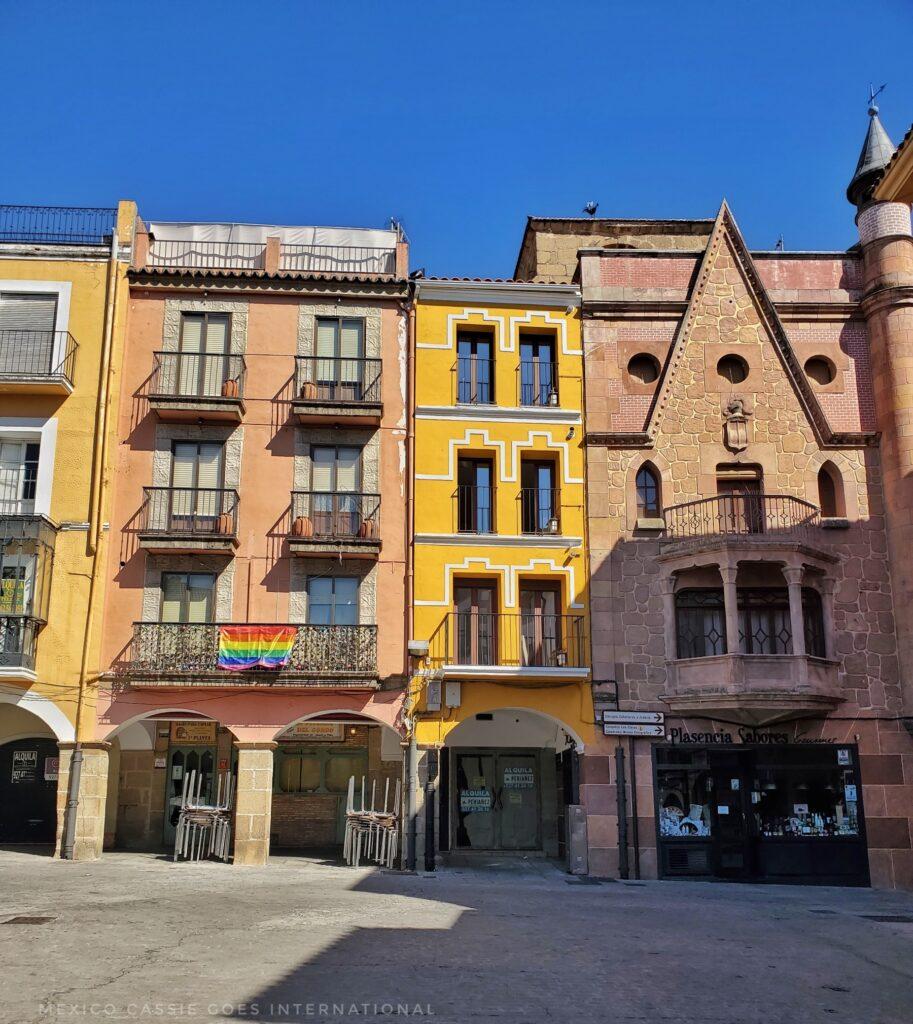 narrow old buildings on a plaza. one on right has pointed 'arch' above a window. one on left has pride flag hanging