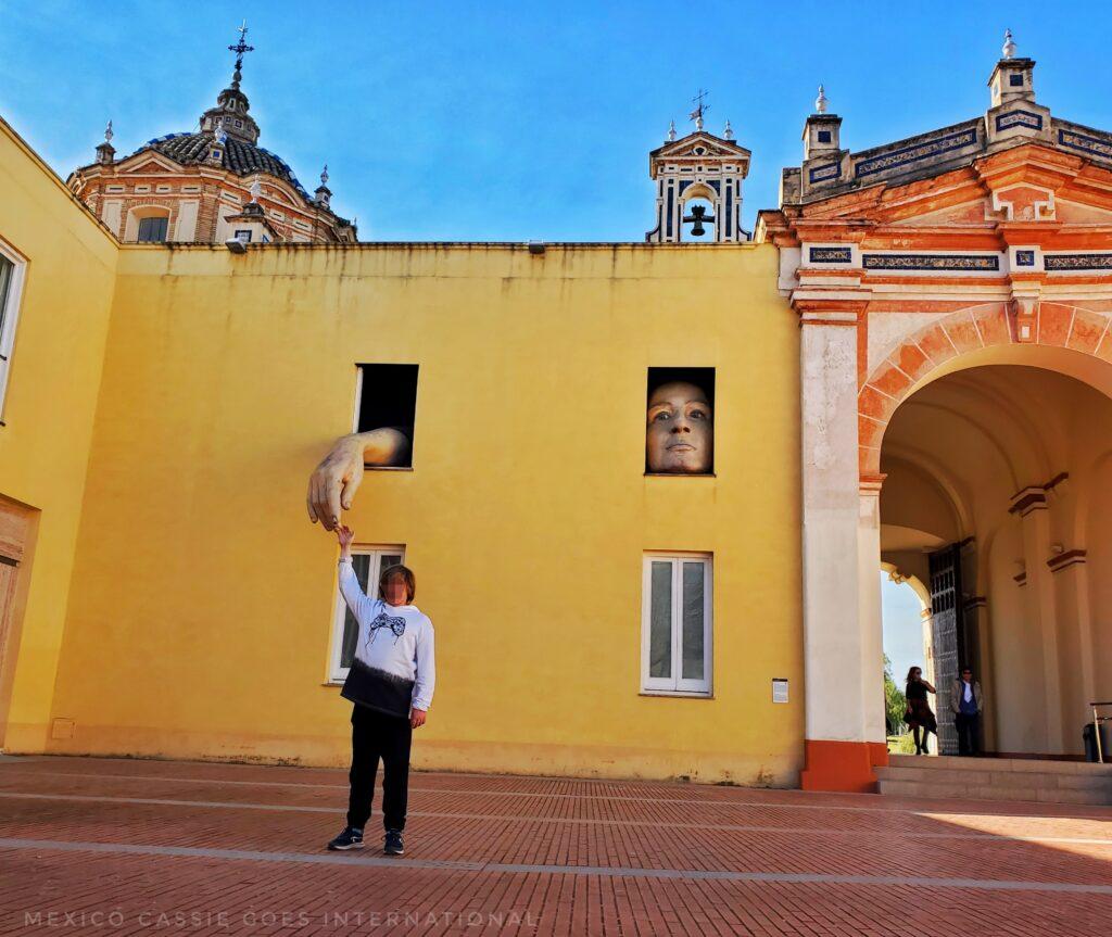 kid standing in front of yellow wall, top windows have a sculpture of a hand and a face, hand is coming out towards the kids