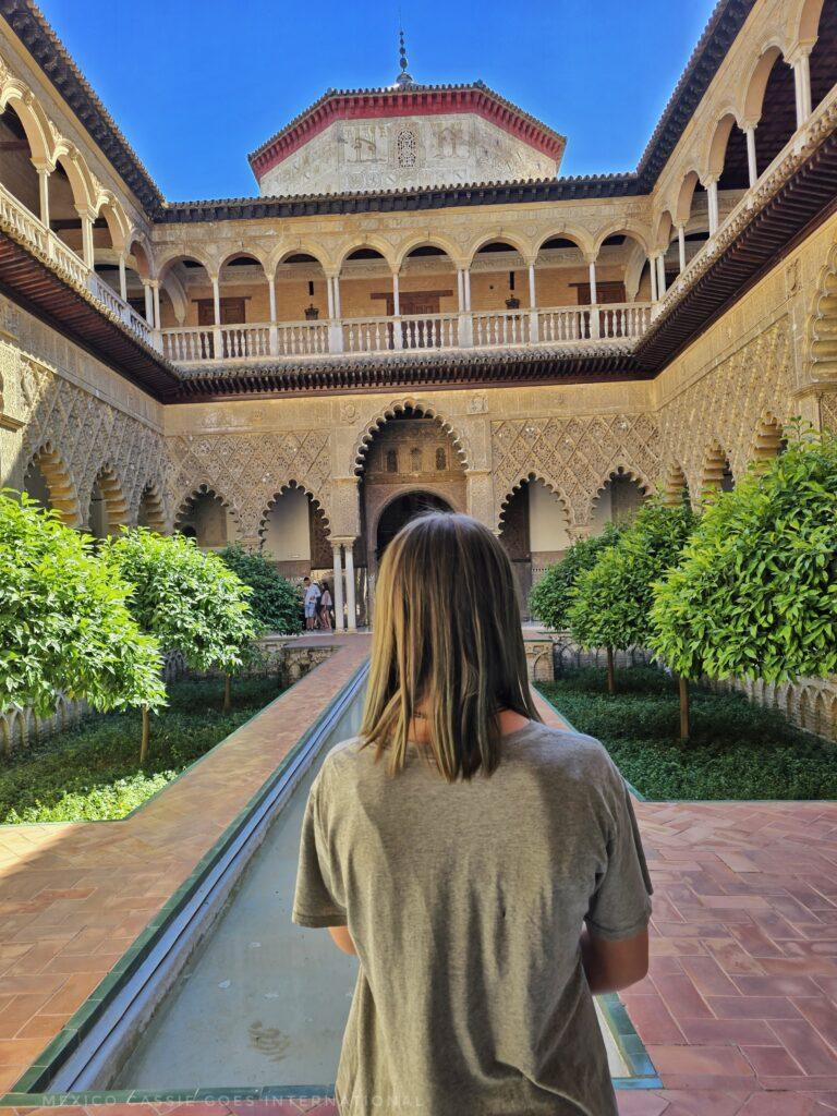 kid in grey tshirt with shoulder length hair standing facing away from the camera in the courtyard of the alcazar (water channel in front, building with arches all around, low green bushes
