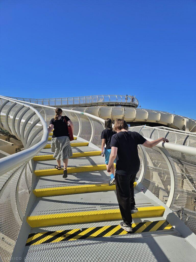 two kids and an adult walking on a walk way with shallow wide steps marked in yellow. pure blue sky