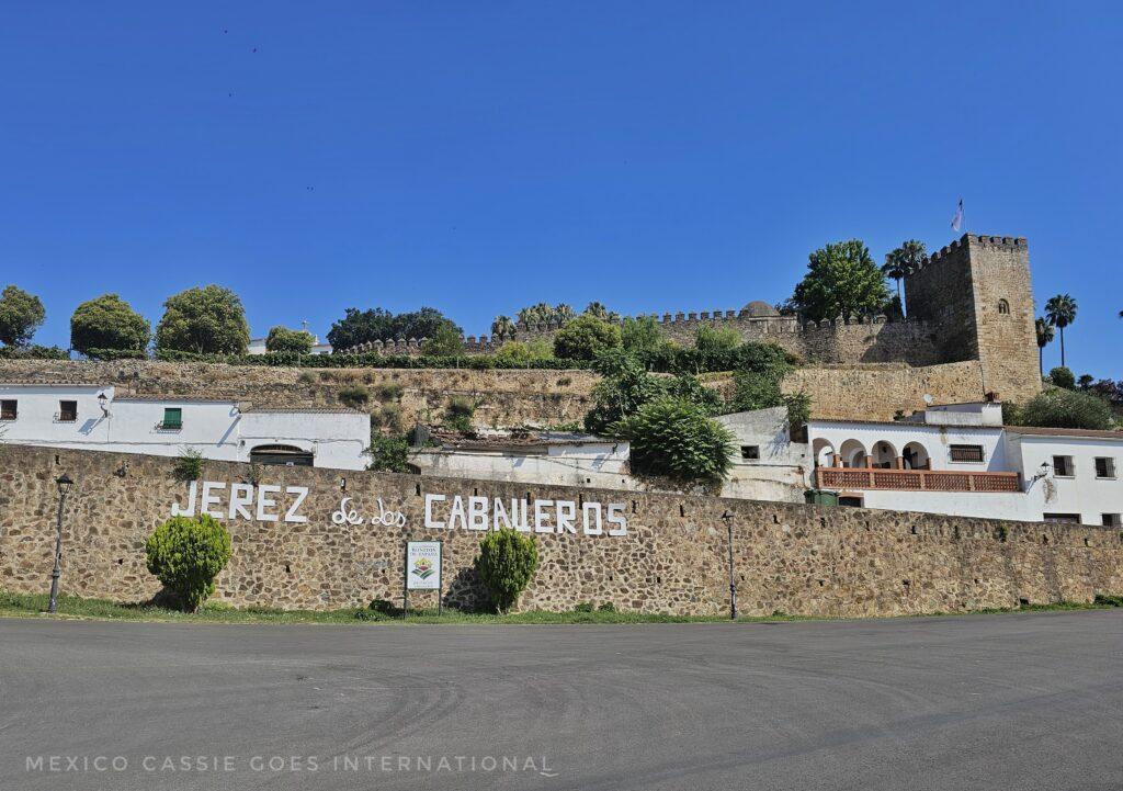 stone wall with jerez de los caballeros painted in white. castle and white buildings above, blue sky and trees
