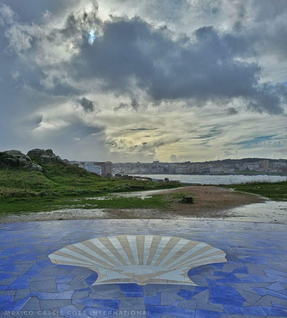 the camino conch shell in tile form on ground, cean, city and sky behind
