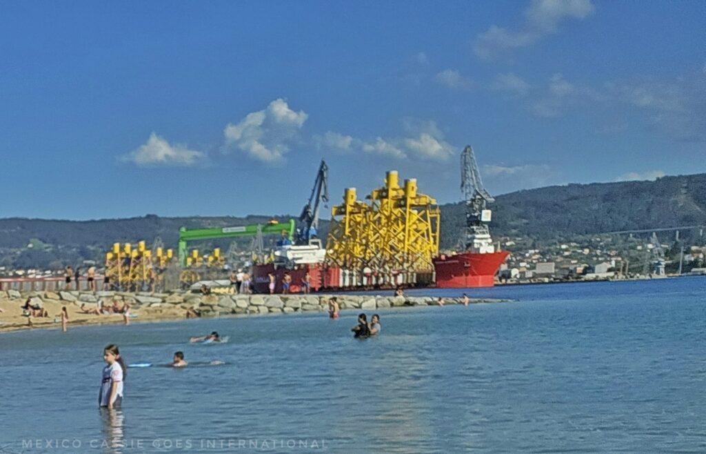 view from beach, people in very calm water, docks in background with yellow crane structures, and red and white structures too