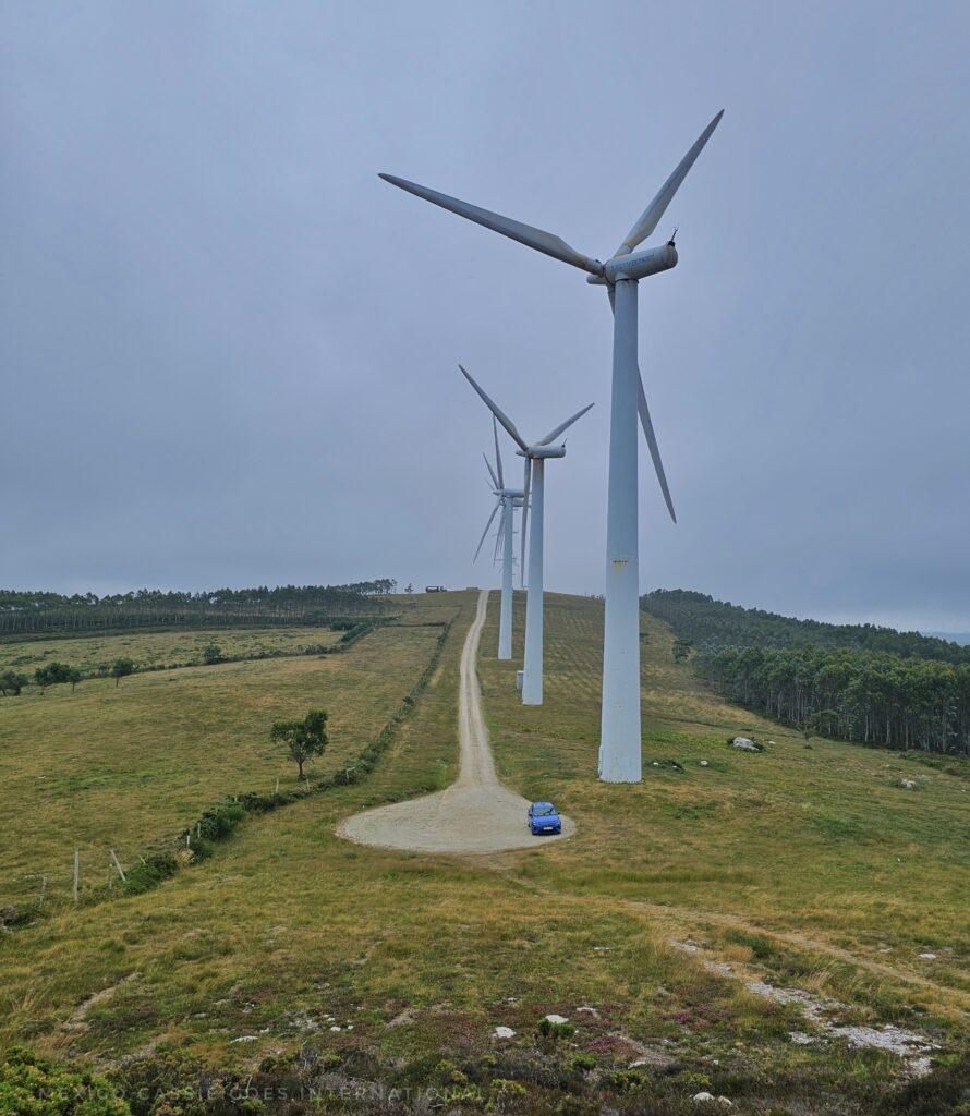 row of wind turbines next to a road, tiny car parked in small circle at end of road