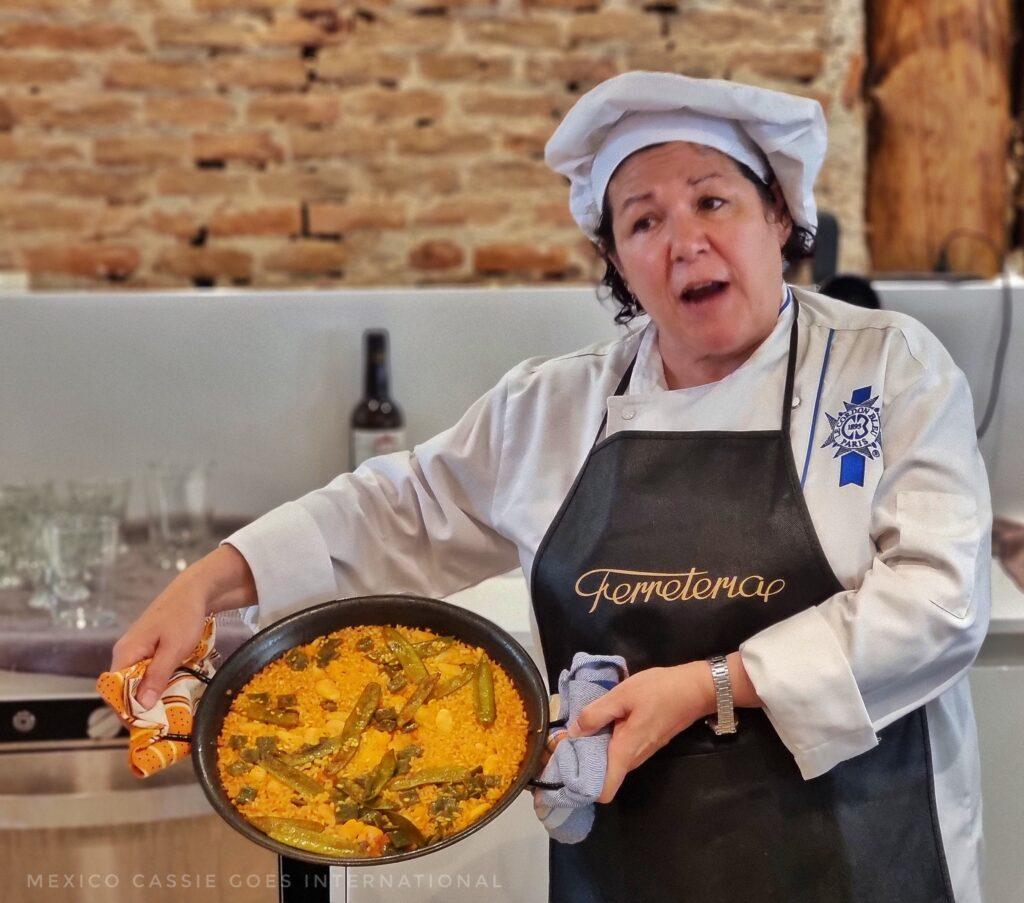 female chef in black apron and white cap holding a cooked paella