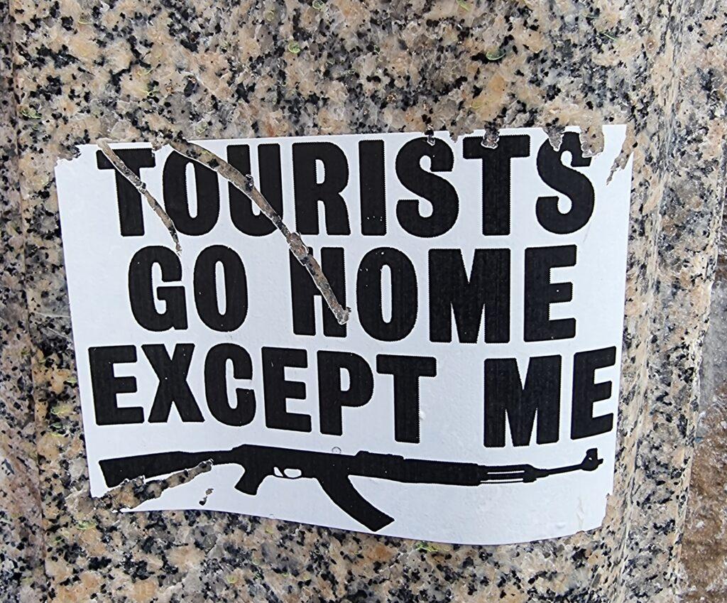 sign in black caps that says "tourists go home except me", gun underneath