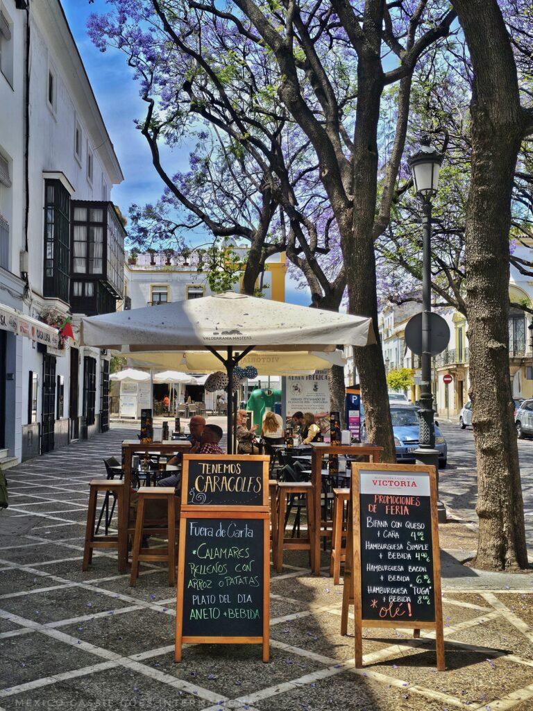outdoor seating at a restaurant. tables have white umbrellas, two chalk boards, trees, building on left