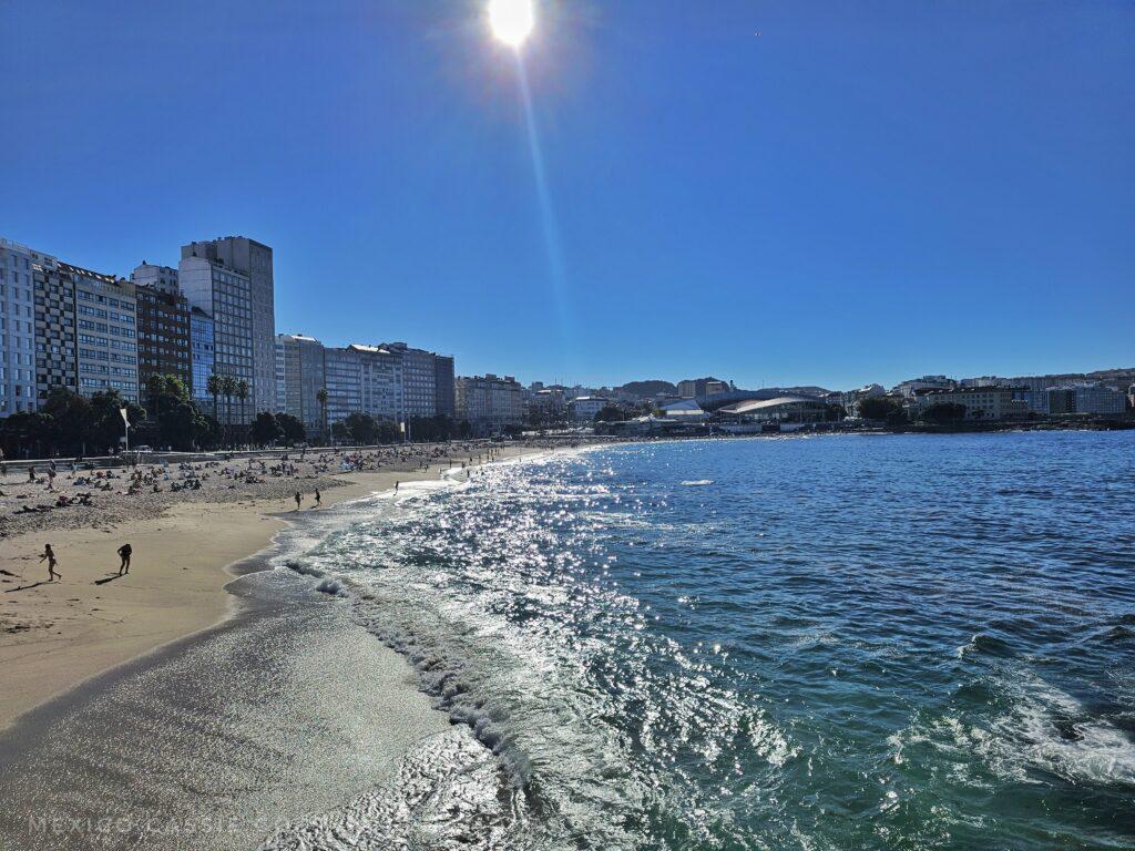 Riazor beach, sun shining, people on beach, blue water, blue sky, buildings along left side